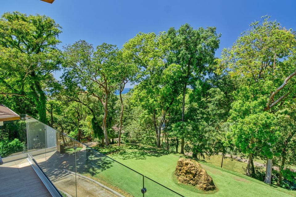 A lush, green outdoor area with a grassy lawn, large trees providing shade, and a prominent rock formation, overlooking a scenic view in Roatan, Honduras.