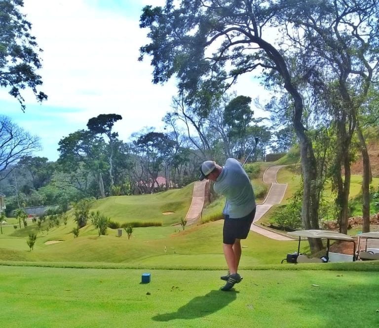 A golfer taking a swing on a lush, green golf course with winding pathways, surrounded by tall trees and natural scenery in Roatan, Honduras.