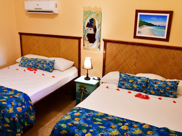 A hotel room in Roatan, Honduras, features two beds with ocean-themed bedding, colorful decorative pillows, a nightstand with a lamp, framed beach artwork, a mirror, and an air conditioning unit on a yellow wall.