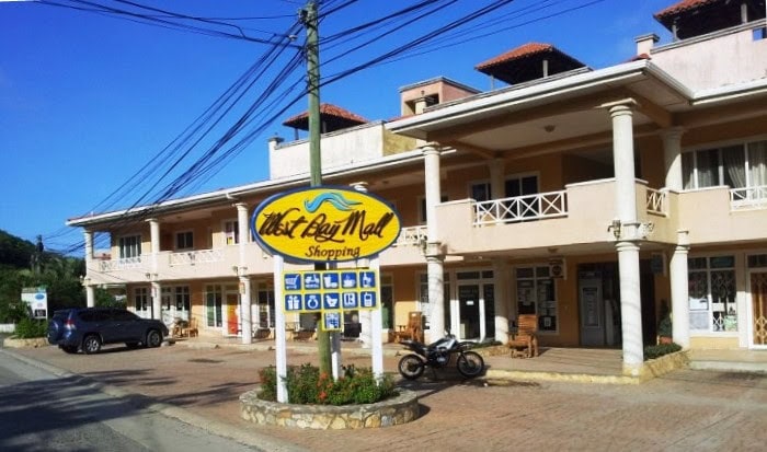 A two-story commercial building in Roatan, Honduras, features a sign for "West Bay Mall Shopping" with icons indicating various shops and services, and parked vehicles including a motorcycle and a car in front of the establishment.