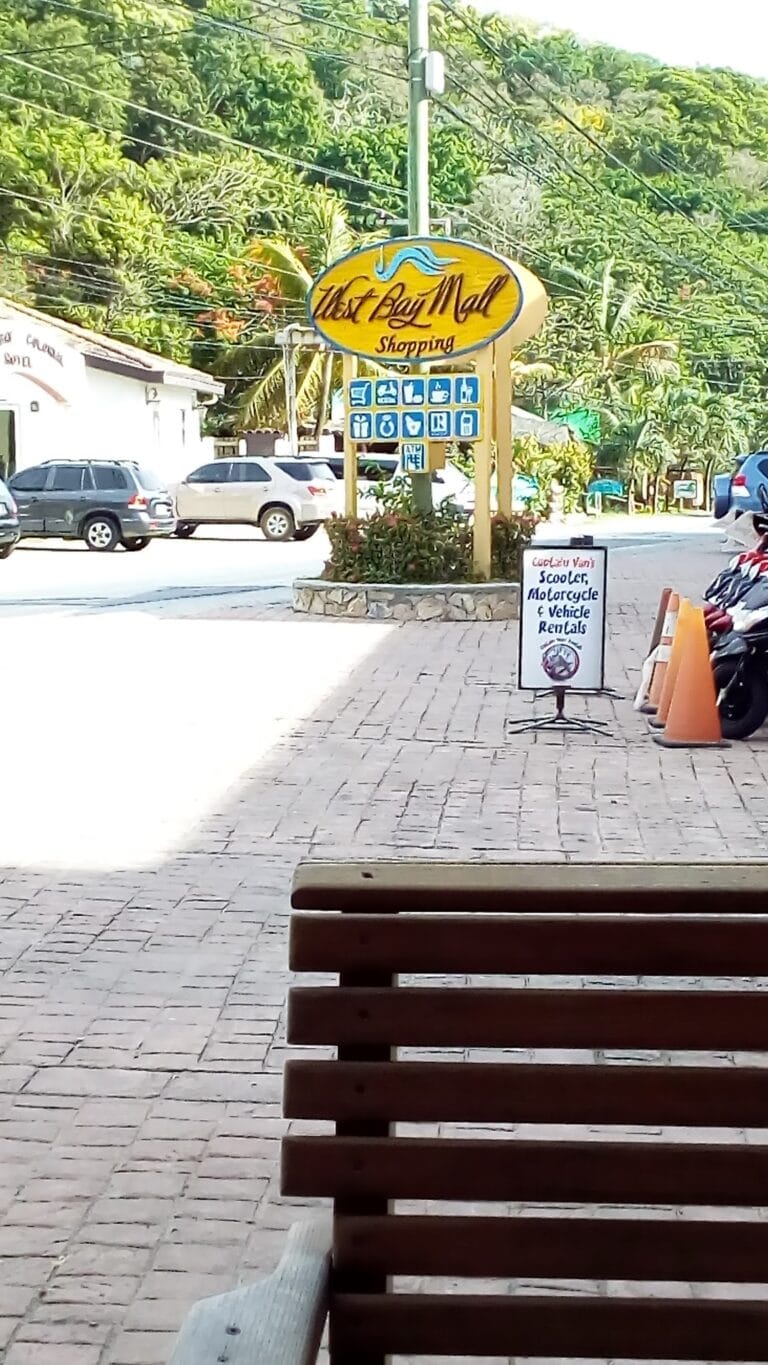 A view from a bench shows a sign for "West Bay Mall Shopping" with parking lot and parked cars, set against lush green trees and utility poles in Roatan, Honduras.