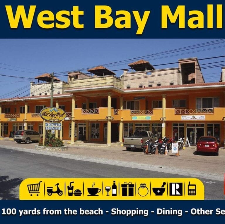 A colorful commercial building with a yellow and orange facade houses shops and businesses at West Bay Mall in Roatan, Honduras, with parked cars, motorcycles, and signage indicating shopping, dining, and services nearby.