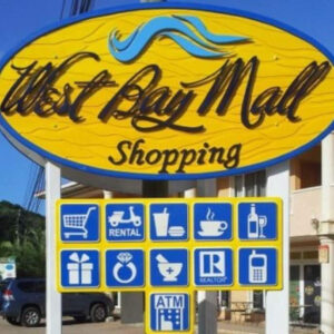 A colorful sign for West Bay Mall Shopping in Roatan, Honduras features icons for shopping, rental services, food and drinks, gifts, jewelry, a pharmacy, real estate, an ATM, and communication services.