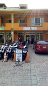 A storefront in Roatan, Honduras offers scooter, motorcycle, and vehicle rentals, with a display of multiple scooters and a red car parked in front, and signage advertising the rental services.