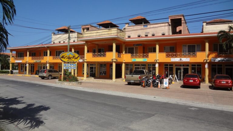 A bright orange commercial building with a sign for "Roatan Art Bay Mall Shopping" in Roatan, Honduras, featuring parking for cars and motorcycles in front under a clear blue sky.
