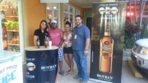 Four people stand around a promotional table and banner for Ron Botran Oro rum outside a store in Roatan, Honduras, with a display of bottles and a backdrop featuring the product branding.