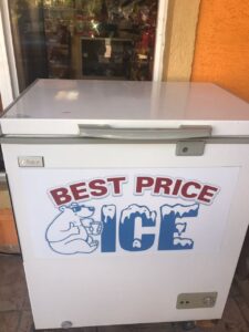 A white chest freezer with a sign that reads "Best Price Ice" and features a cartoon polar bear holding a cup, located outside a building in Roatan, Honduras.