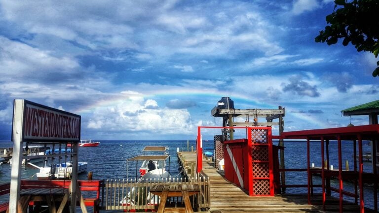 A seaside dock in Roatan, Honduras, features boats moored along the wooden pier with a cloudy sky and a faint rainbow overhead, adjacent to a building with a green roof and a red fence.