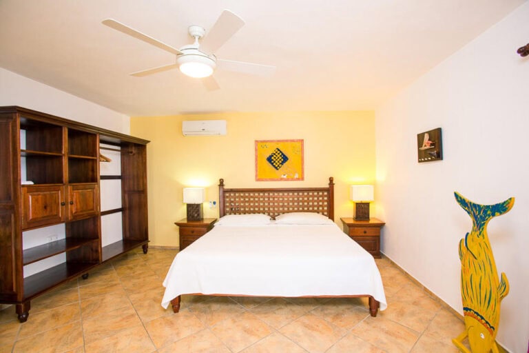 A hotel bedroom in Roatan, Honduras, features a double bed with white bedding, two wooden nightstands with lamps, a yellow accent wall with artwork, a ceiling fan, an air conditioning unit, a large empty wooden wardrobe, and colorful decorative items including a painted whale tail and artwork on the white walls.