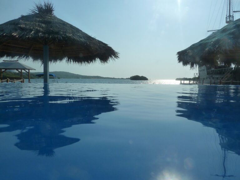 A view of a pool with thatched-roof palapas, overlooking calm waters and distant islands on Roatan, Honduras, under a bright, sunny sky.