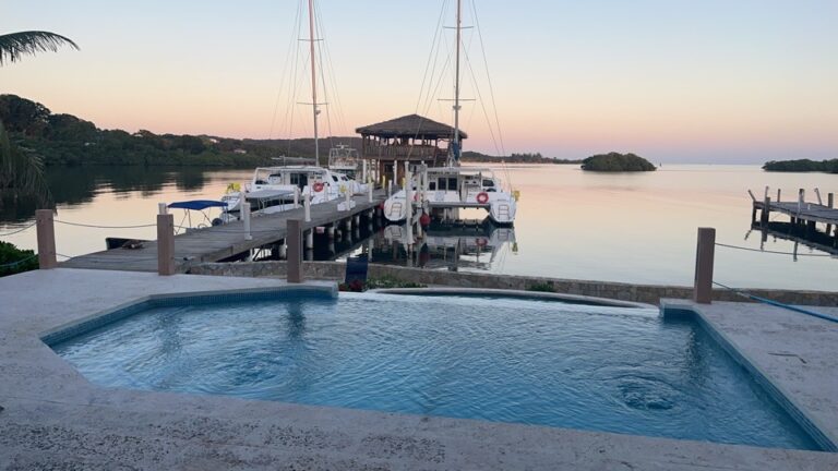 A swimming pool overlooking a calm waterside marina with sailboats and luxury yachts in Roatan, Honduras, during sunset.