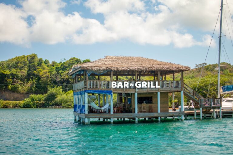 A wooden bar and grill with a thatched roof is built on stilts over the turquoise waters of Roatan, Honduras, featuring an outdoor seating area, hammocks, and scenic views of lush green hills and boats docked nearby.