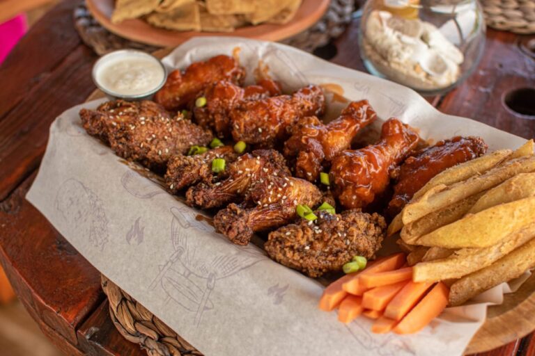 A platter of assorted fried and sauced chicken wings, potato wedges, carrot sticks, and green onions served with dipping sauces on a wooden table in Roatan, Honduras.