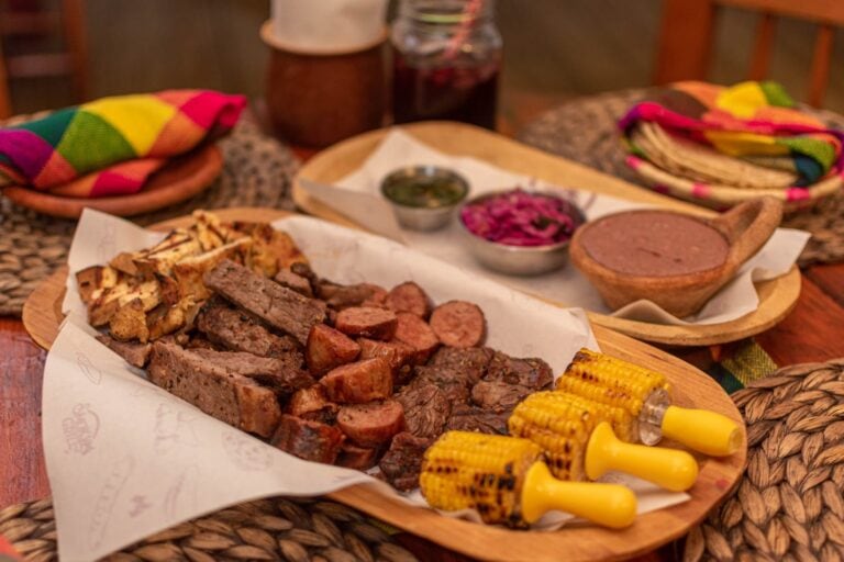 A traditional Honduran meal featuring grilled meats, corn on the cob, and various side dishes is served on a wooden tray at a rustic table in Roatan, Honduras, with colorful woven cloth napkins in the background.
