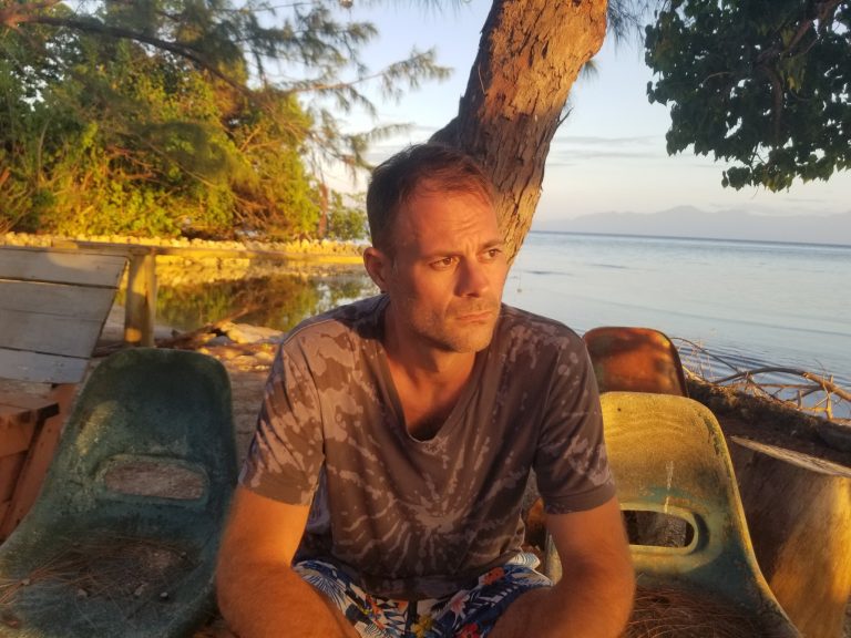 A man in camouflage shirt and patterned shorts sits on a stone bench near the beach in Roatan, Honduras, with lush green trees, a wooden railing, and calm ocean waters backdropped by a clear sky during sunset.