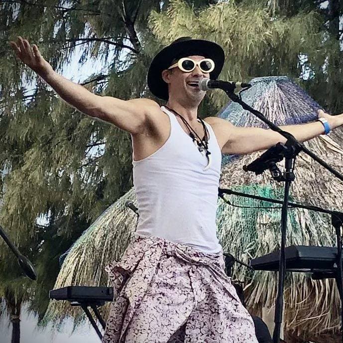 A person wearing a black wide-brimmed hat, white sunglasses, a white tank top, and patterned pants performs on an outdoor stage in Roatan, Honduras, with arms outstretched and a microphone in front of a palm tree and tropical foliage.
