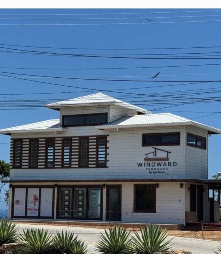 A modern two-story building with white exterior walls and dark window frames, labeled "Windward Terraces by Sgt Rico's," located in Roatan, Honduras, features large windows, a wooden balcony on the upper level, and is surrounded by desert-style plants beneath a clear blue sky with electrical wires and birds overhead.