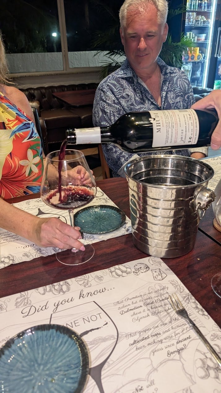 A person in a floral shirt is pouring red wine from a bottle into a glass at a restaurant in Roatan, Honduras, with a man sitting nearby and a wine refrigerator in the background.