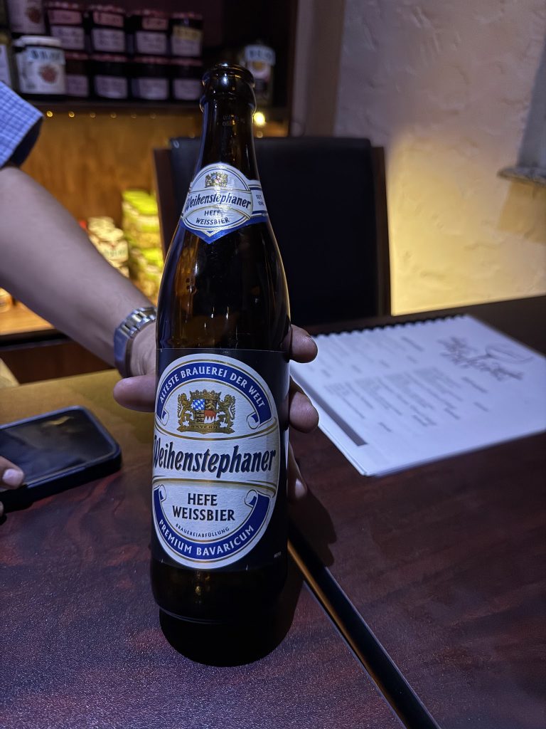 A person holds a bottle of Weihenstephaner Hefe Weissbier in front of a restaurant or bar setting in Roatan, Honduras, with a menu on the table and shelves of food items in the background.