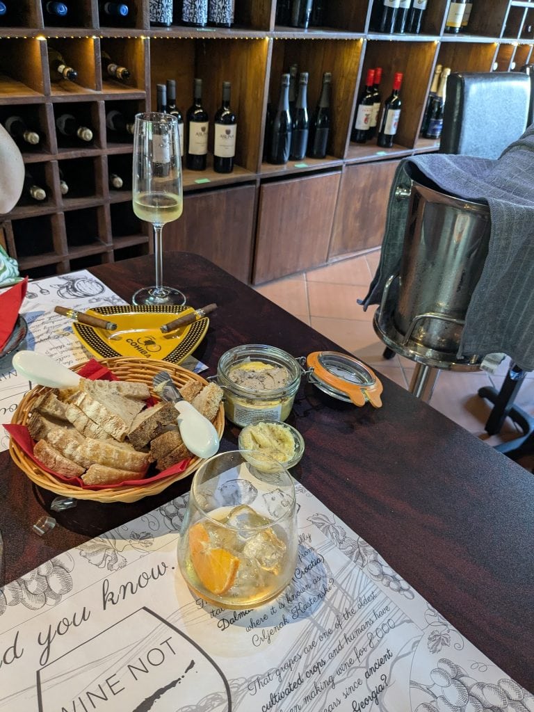 A dining table in a Roatan, Honduras restaurant features a basket of sliced bread, small dishes of dips, a glass of iced lemonade with lemon and orange slices, and a menu with decorative floral borders, with a wooden wine storage shelf in the background.