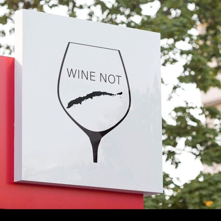 A sign featuring a wine glass with the words "Wine Not" inside, displayed outdoors against a background of green trees on Roatan, Honduras.