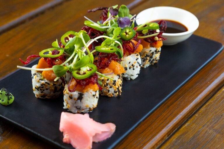 A plate of sushi topped with microgreens, sliced vegetables, and black sesame seeds is presented with soy sauce and pickled ginger on a wooden table in Roatan, Honduras.