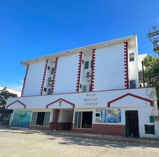 The Wood Medical Center building in Roatan, Bay Islands, Honduras, is a white multi-story structure with red accents and signages, under a clear blue sky.