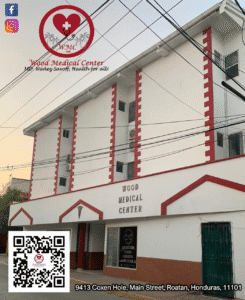 The image shows a white multi-story building with red decorative accents and signage for Wood Medical Center located on Main Street in Roatan, Honduras.