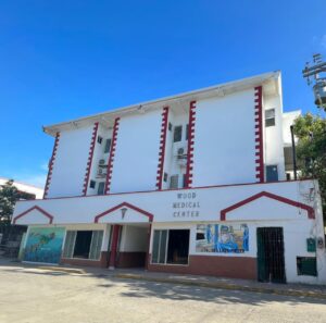 The Wood Medical Center building in Roatan, Honduras, features a white facade with red vertical accents, signage indicating it is a medical facility, and a mural depicting healthcare workers wearing masks on the lower portion of the structure under a clear blue sky.