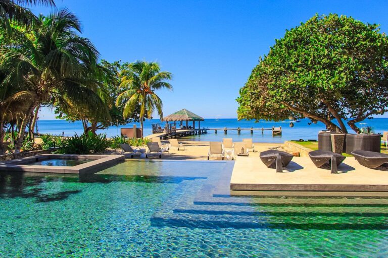 A luxurious beachfront resort in Roatan, Honduras features an infinity swimming pool overlooking the ocean, surrounded by palm trees, lounge chairs, and a shaded gazebo on a sandy beach with a clear blue sky and calm sea in the background.