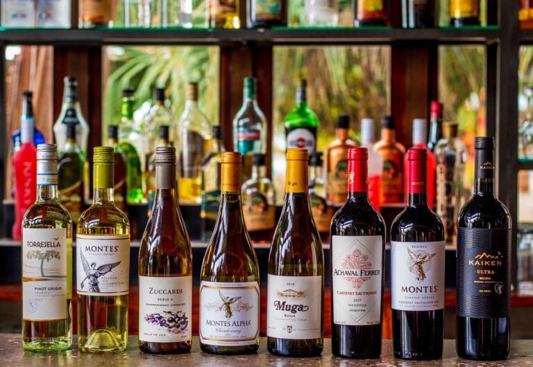 A selection of local and international wine bottles displayed on a bar counter in Roatan, Honduras, with a wooden shelf of various liquor bottles in the background and lush greenery visible through the windows.