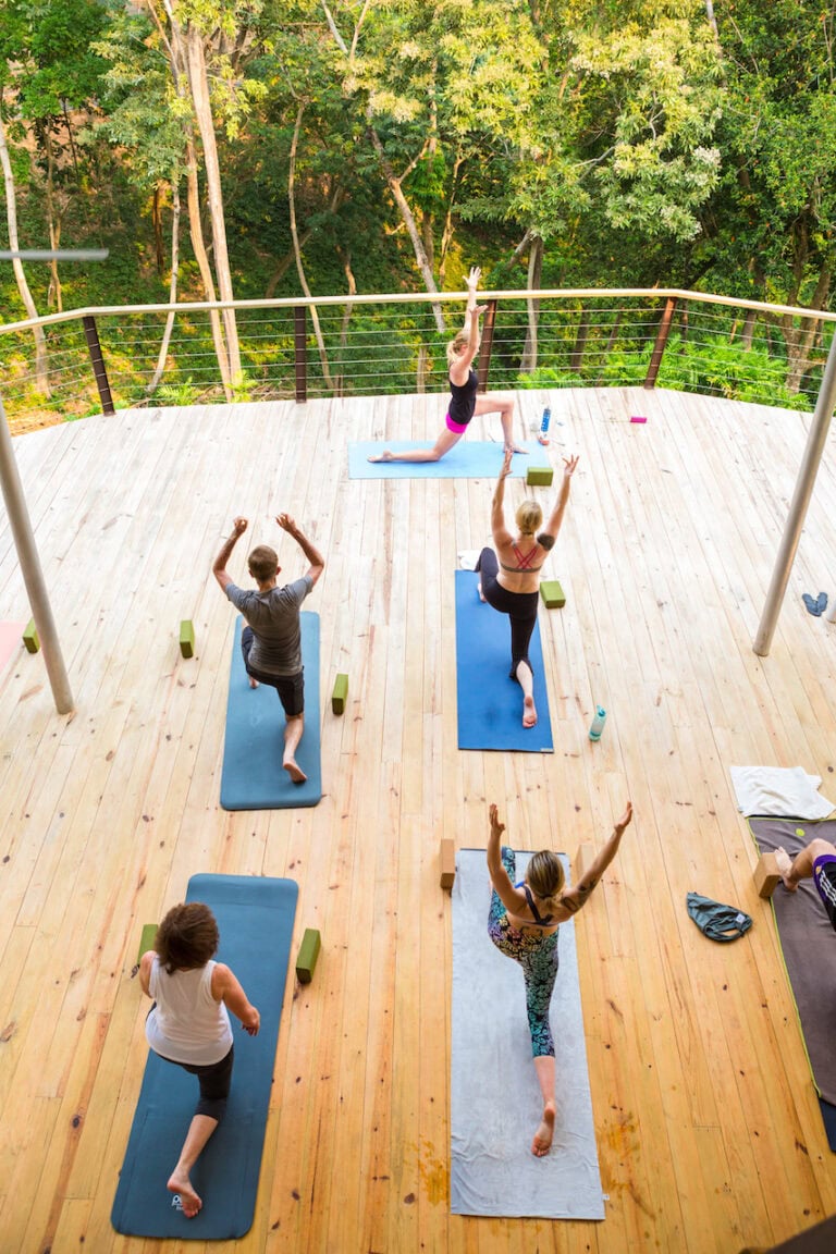 A group of people practicing yoga on wooden outdoor decking surrounded by trees in Roatan, Honduras.