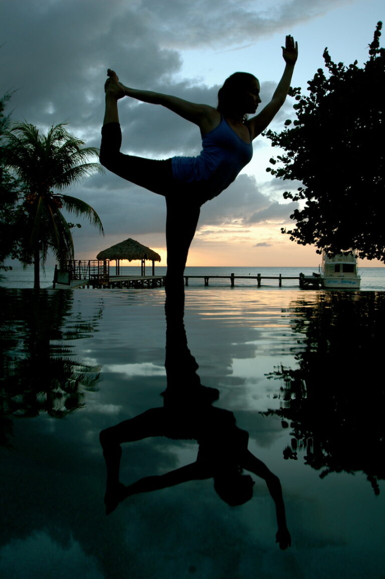 A woman practicing yoga in the dancer pose beside a reflective pool at sunset in Roatan, Honduras, with a palm tree, a thatched-roof hut, and a boat on the ocean in the background.