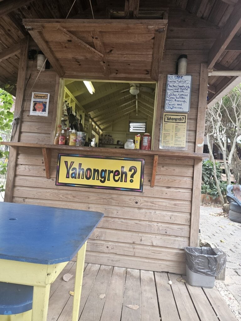 A small wooden food stand near the beach in Roatan, Honduras, features a colorful sign reading "Yahongreh?" and handwritten menu boards listing various food and drink options.