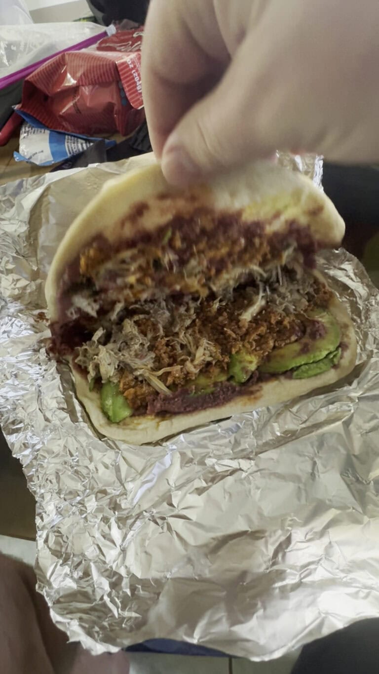 A partially assembled shredded beef taco with guacamole in a soft flour tortilla, resting on crumpled aluminum foil, with a blurred background including snacks and a hand holding the taco, on a table in Roatan, Honduras.