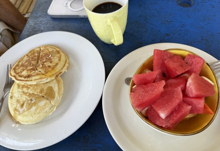 A breakfast scene on Roatan, Honduras, showing two plates with pancakes and watermelon, a mug of coffee, and a digital scale on a blue table.