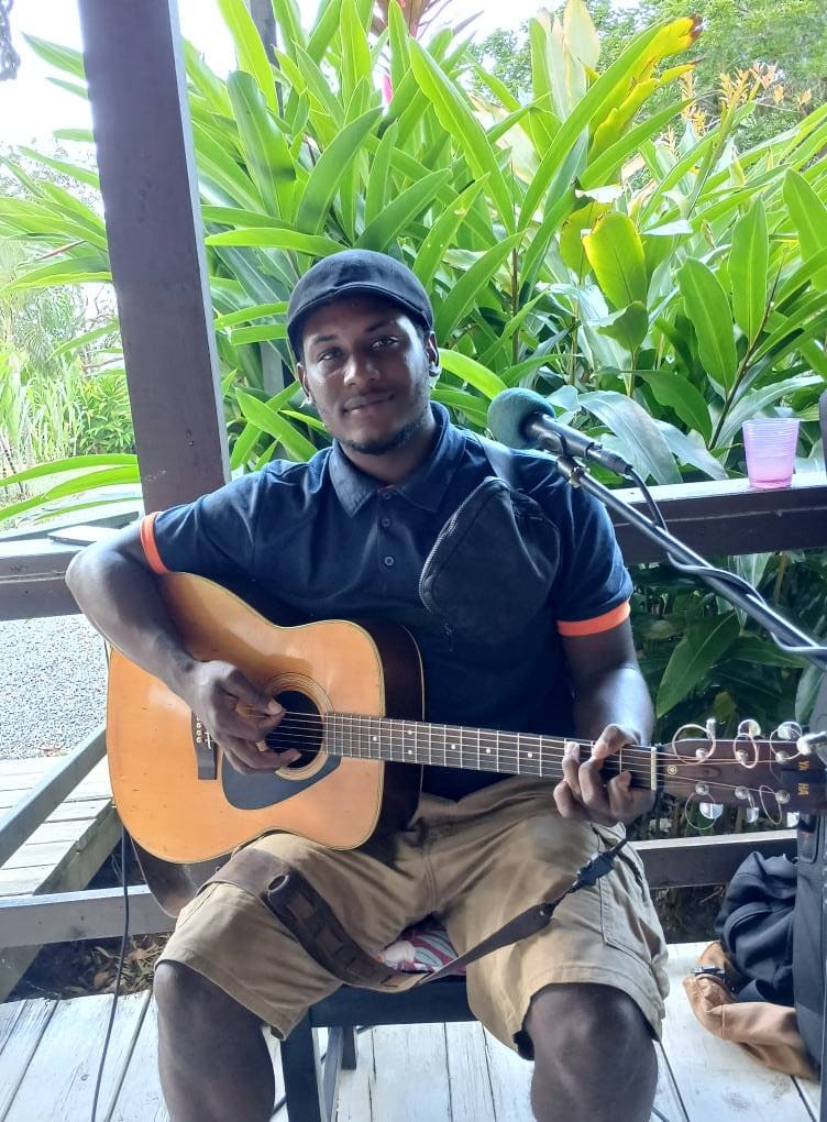 A man wearing a black cap and navy blue shirt plays an acoustic guitar and sings into a microphone on a shaded outdoor deck, with lush green tropical plants and trees in the background, in Roatan, Honduras.